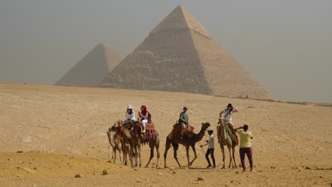 Tourists-sit-on-top-of-camels-in-the-desert-with-great-pyramids-in-the-background