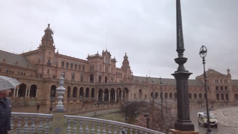 Tourists-with-umbrellas-at-rainy-Plaza-de-Espana-in-Seville,-Spain,-pan-right