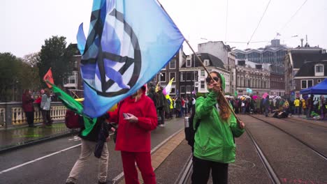 Two-Girls-Waving-Flags-With-Group-Of-Protesters-In-The-Back-At-Blauwbrug-Bridge-During-The-The-Extinction-Rebellion-Climate-Protest-In-Amsterdam,-Netherland