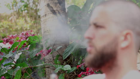 Green-vegetation-and-red-flowers-in-the-background,-in-front-of-a-defocused-bearded-bald-man-smoking-vape