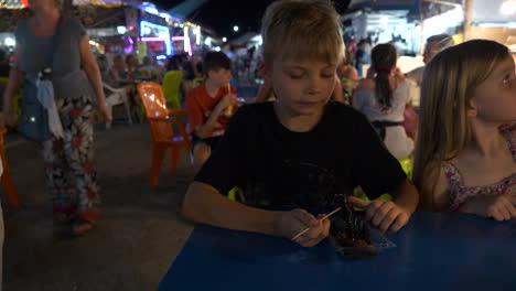 Boy-eating-a-scorpion,-accompanied-by-family,-at-a-local-market,-in-Khao-Lak,-Thailand