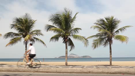 People-with-and-without-face-masks-running,-jogging,-biking,-walking,-cycling-and-on-skateboard-passing-three-palm-trees-and-Ipanema-beach-during-COVID-19-coronavirus-outbreak
