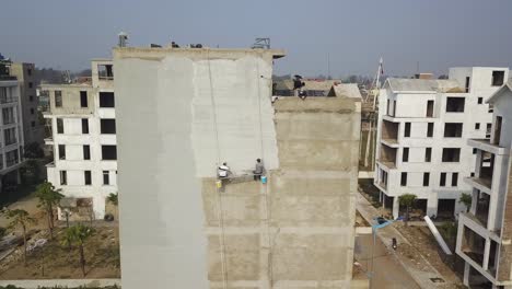 Construction-workers-swinging-on-the-side-of-a-building-applying-concrete-plaster-finish,-Aerial-pedestal-lift-shot