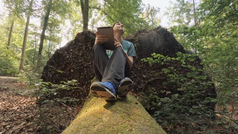 A-man-is-working-at-his-tablet-while-he's-sitting-at-a-mossy-trunk-in-a-green-forest---business-working-at-the-fresh-air,-modern-technology