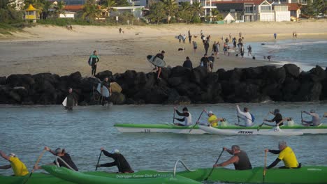 Groups-Of-Creek-Paddlers-Racing-In-Currumbin-Beach---Surfers-Passing-Over-The-Rocks-Into-The-Sea-On-A-Sunset---Water-Activities-In-Gold-Coast,-Queensland---wide-slowmo-shot