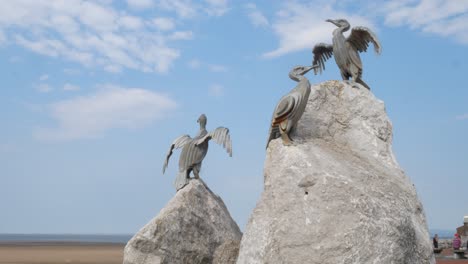 Stone-resort-jetty-promenade-cormorant-bird-sculptures-at-Morecambe-beach-under-blue-sky-wide-left-dolly
