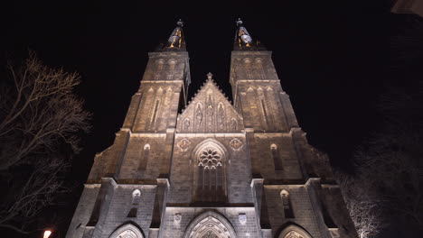 Der-Beleuchtete-Eingang-Und-Die-Vorderseite-Der-Gotischen-Mittelalterlichen-Steinkirche-St.-Peter-Und-Paul-Mit-Ihren-Beiden-Türmen-In-Vyšehrad,-Prag,-Tschechien,-Nachts,-Auf-Einem-Leeren-Gepflasterten-Platz-Mit-Laternen,-Geneigt
