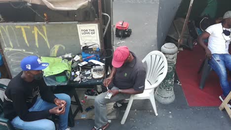 An-informal-street-market-area-showing-a-black-man-making-repairs-to-a-mobile-device-with-MTN-branding-painted-on-his-street-market-vendor-stall