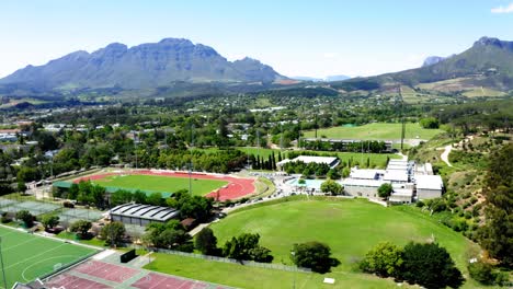 Aerial-drone-rotating,-university-college-sports-athletics-stadium,-track-and-field,-tennis-courts,-olympic-swimming-pool,-mountains-and-trees-in-background,-Stellenbosch,-Coetzenburg