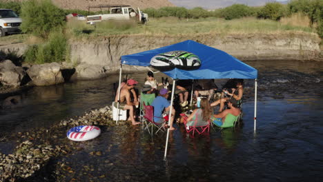 Group-of-young-people-sitting-in-calm-river-celebrating-4th-of-July-under-tiny-party-tent