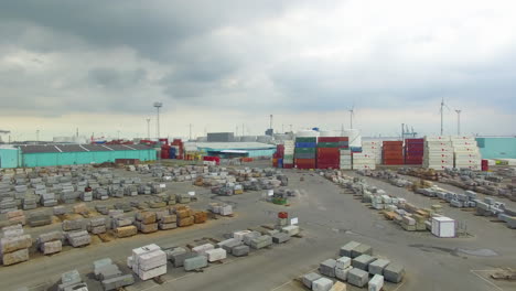 Aerial-footage-near-the-Scheldt-river-in-the-port-of-Antwerp,-cargo-ship,-machinery,-granite-blocks,-containers,-industry-area-with-many-windmills-in-the-horizon
