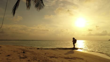 Backpacker-standing-over-big-rock-on-exotic-beach-under-a-palm-tree-at-sunset-moments-with-yellow-sky-reflecting-on-calm-sea-in-Philippines