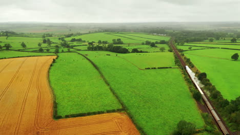 Aerial-shot-following-a-steam-train-with-white-smoke-traveling-through-the-English-countryside,-cloudy-day