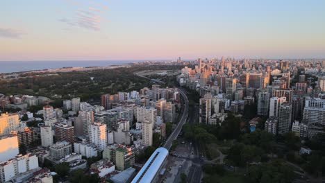 Dolly-in-flying-over-a-train-station-surrounded-by-buildings-and-parks-with-Rio-de-la-Plata-river-in-background,-Buenos-Aires,-Argentina