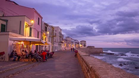 Scenic-timelapse-of-people-walking-by-city-walls-and-the-ocean-in-the-Alghero-town,-Sardinia,-with-a-beautiful-small-cafe-in-the-foreground