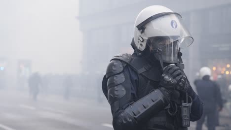A-single-police-officer-in-full-riot-gear-places-a-gas-mask-on-his-face-during-a-smokey-riot,-slow-motion