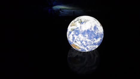 Luke-Jarram-Floating-Earth-illuminated-planet-reflecting-in-lake-water-rippling-surface-at-night-rising-pull-back