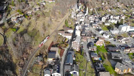 Aerial:-red-and-white-train-in-the-Alps,-going-along-a-mountainside-and-leaving-an-alpine-village
