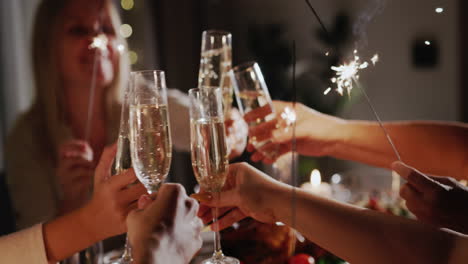A-group-of-people-at-the-festive-table-clink-glasses,-holding-sparklers-in-their-hands.-New-Year's-Eve