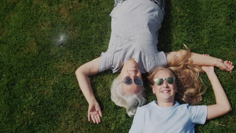 Senior-woman-and-granddaughter-lie-on-the-grass-under-jets-of-water,-laugh-and-escape-from-the-heat.-Slow-motion-video