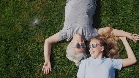 Grandmother-and-granddaughter-lie-on-the-grass-under-jets-of-water,-laugh-and-escape-from-the-heat.-Slow-motion-video