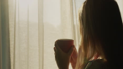 Back-view:-A-woman-with-a-cup-of-hot-tea-looks-out-the-window-where-the-sun-has-risen.-Hot-drink-and-good-mood