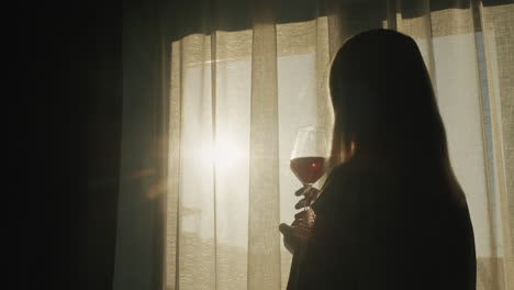 A-woman-with-a-glass-of-red-wine-stands-at-the-window-at-sunset
