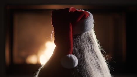 A-girl-with-long-blond-hair-in-a-New-Year's-cap-looks-at-the-fire-in-the-fireplace.-Back-view