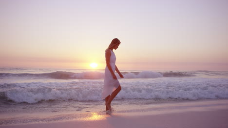 beautiful-woman-wearing-white-dress-walking-along-shore-line-on--beach-at-sunset-in-slow-motion-RED-DRAGON