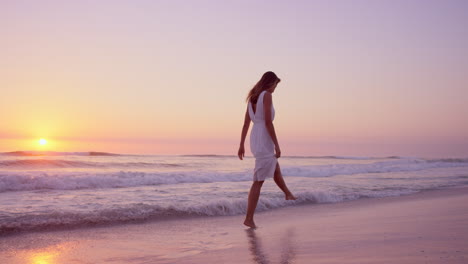 beautiful-woman-wearing-white-dress-walking-along-shore-line-on--beach-at-sunset-in-slow-motion-RED-DRAGON