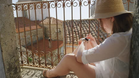 Mujer-Usando-Un-Reloj-Inteligente-Revisando-Mensajes-Navegando-Disfrutando-De-Un-Cálido-Día-Soleado-De-Vacaciones-Sentado-En-El-Balcón-Turista-Feliz-Relajándose-Con-Sombrero-En-Italia