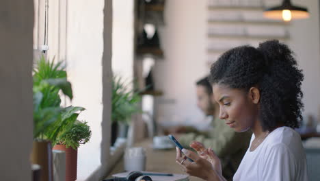 Feliz-Mujer-Afroamericana-Usando-Un-Teléfono-Inteligente-En-Una-Cafetería-Navegando-Mensajes-En-Línea-Disfrutando-De-Compartir-El-Estilo-De-Vida-En-Las-Redes-Sociales-Relajándose-En-Un-Restaurante-Cafetería