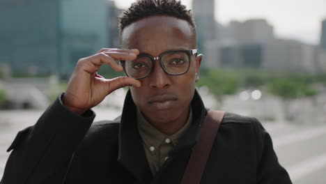 close-up-portrait-of-young-trendy-african-american-man-student-puts-on-glasses-looking-at-camera-smiling-confident-in-urban-city-background