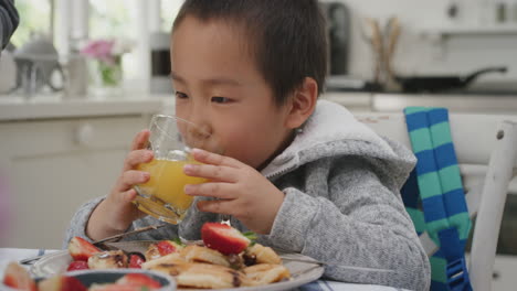 Lindo-Niño-Asiático-Bebiendo-Jugo-Comiendo-Gofres-Para-El-Desayuno-Disfrutando-De-Una-Comida-Casera-Saludable-Con-La-Familia-En-La-Cocina-De-Casa-4k
