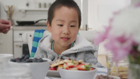 Lindo-Niño-Asiático-Comiendo-Gofres-Frescos-Para-El-Desayuno-Disfrutando-De-Una-Deliciosa-Comida-Casera-Con-La-Familia-En-La-Cocina-De-Casa-4k