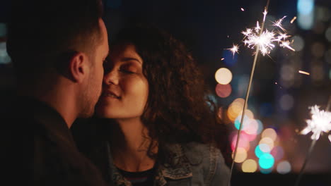 happy-caucasian-couple-kissing-on-rooftop-at-night-holding-sparklers-celebrating-anniversary-enjoying-romantic-urban-evening