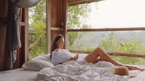 beautiful-woman-using-smartphone-in-bed-at-hotel-resort-texting-sharing-vacation-relaxing-in-comfort-with-view-of-tropical-jungle
