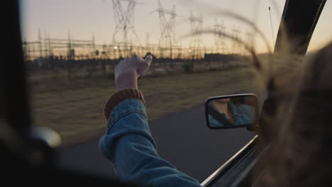 young-woman-in-car-holding-hand-out-window-feeling-wind-blowing-through-fingers-driving-in-countryside-on-road-trip-travelling-for-summer-vacation-enjoying-freedom-on-the-road-at-sunset