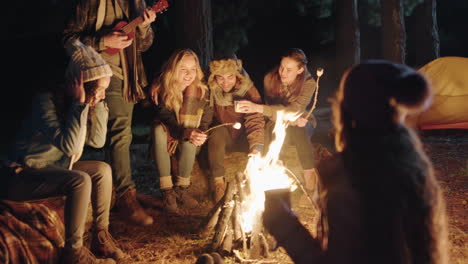 happy-friends-sitting-by-campfire-young-man-playing-ukelele-singing-enjoying-evening-together-camping-in-forest-at-night