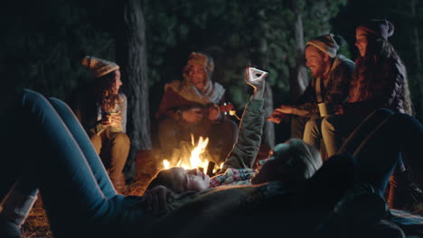 camping-friends-using-smartphone-browsing-social-media-lying-on-ground-by-campfire-at-night-in-forest