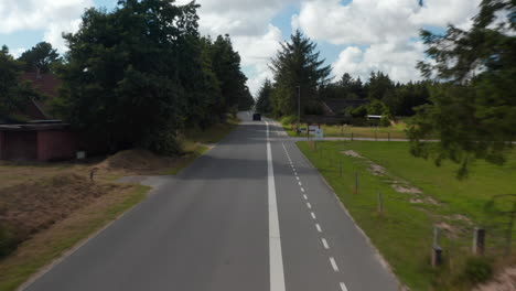 Forwards-fly-above-road-with-cyclist-path-leading-through-countryside-with-trees-and-houses.-Car-driving-on-road.-Denmark