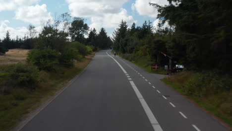 Low-flight-above-road,-passing-by-cyclist-riding-on-cyclist-path.-Ascending-and-revealing-wooded-flat-landscape.-Denmark