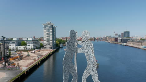 Backwards-reveal-of-Molecule-Man-sculpture-in-Spree-river-and-various-buildings-surrounding-calm-water.-Sunny-day-with-clear-sky.-Berlin,-Germany