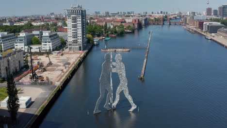 Forwards-descending-fly-towards-Molecule-Man-near-bank-of-Spree-river.-Sculpture-of-three-human-silhouettes-from-irregularly-perforated-material.-Berlin,-Germany