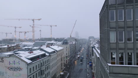 Fly-above-streets-in-urban-neighbourhood-in-winter.-Group-of-tower-cranes-on-construction-site.-Falling-snow-covering-multistorey-building-roofs.-Berlin,-Germany