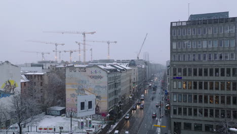 Elevated-footage-of-buildings-around-street-intersection-in-urban-neighbourhood.-Group-of-tower-cranes-on-construction-site-in-background.-Winter-weather-with-snowing.-Berlin,-Germany