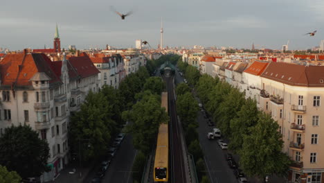 Backwards-tracking-scan-train-driving-in-wide-street-with-trees.-Surrounding-buildings-illuminated-by-bright-morning-sun.-Fernsehturm-TV-tower-on-horizon.-Berlin,-Germany