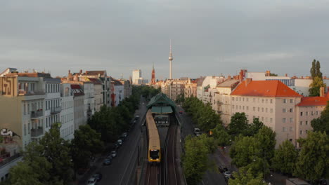 Backwards-tracking-of-Sbahn-train-leaving-train-stop-above-wide-street-in-city.-Morning-view-of-urban-neighbourhood.-Fernsehturm-TV-tower-in-background.-Berlin,-Germany