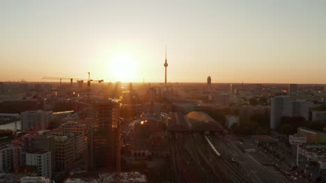 AERIAL:-Berlin,-Germany-construction-site-with-cranes-with-beautiful-sunset,-sunlight-and-view-on--Alexanderplatz-TV-Tower,-Sunflairs