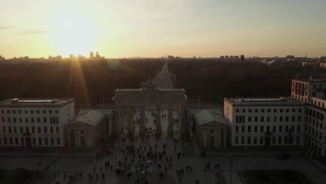 AERIAL:-Brandenburg-Gate-in-Berlin-with-crowd-of-people,-tourists-on-the-ground-in-beautiful-sunset-light